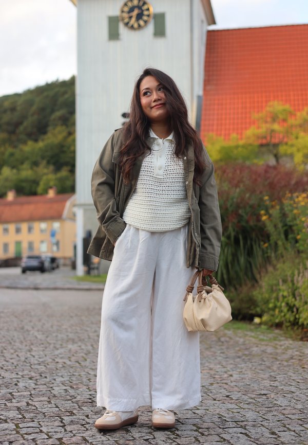 Olive green jacket over a white crochet top, paired with wide white trousers and beige handbag; cream shoes with brown accents.