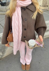 Hnědý vlněný kabát s velkými černými knoflíky, světle růžový pletený šál, světle růžové kalhoty, hnědé platformové boty, držící hrnek s kávou a náčiní.
