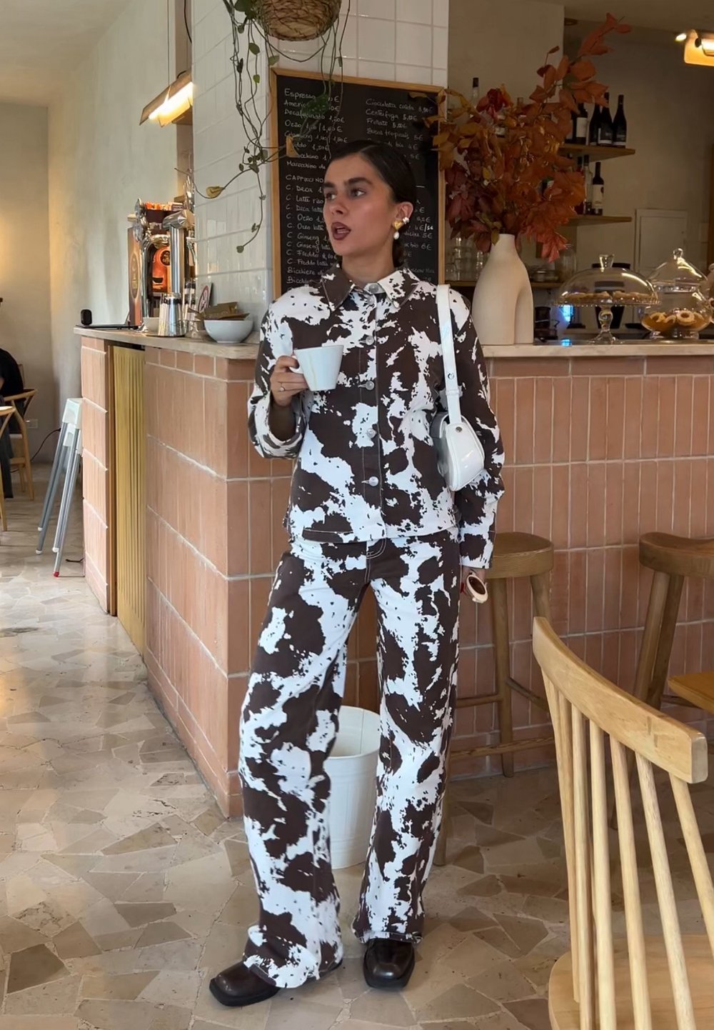 Casaco e calças com padrão de vaca castanho e branco, combinados com sapatos pretos. O conjunto apresenta um ajuste descontraído e cores contrastantes, segurando um copo branco.