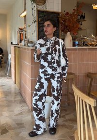Casaco e calças com padrão de vaca castanho e branco, combinados com sapatos pretos. O conjunto apresenta um ajuste descontraído e cores contrastantes, segurando um copo branco.