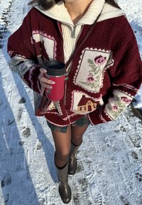 Personne portant un cardigan en maille bordeaux à motifs floraux, une jupe à carreaux et des bottes, tenant un mug de voyage rouge en marchant sur un sol enneigé.