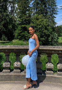 Robe bleu clair sans bretelles avec un corsage ajusté et un ourlet évasé, associée à des sandales à talons transparents ornées de décorations blanches. Tient un ballon.