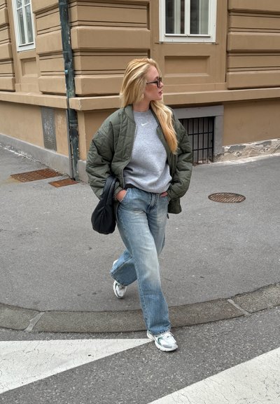 Chaqueta bomber acolchada verde, sudadera gris con logo, jeans de pierna ancha azul claro, bolso negro y zapatillas blancas, caminando por una calle.