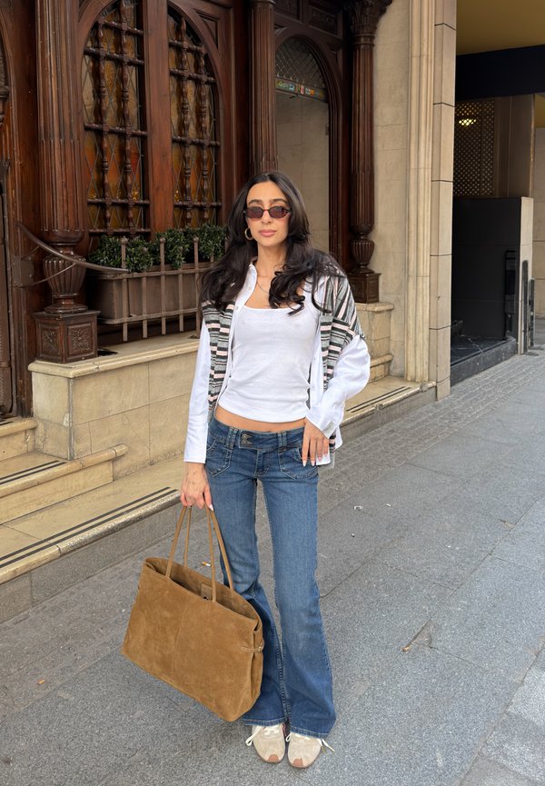 Vrouw met lang donker haar draagt een zonnebril, een wit shirt, een gestreepte cardigan, blauwe jeans, witte sneakers en draagt een grote bruine tas op de stoep in de stad.