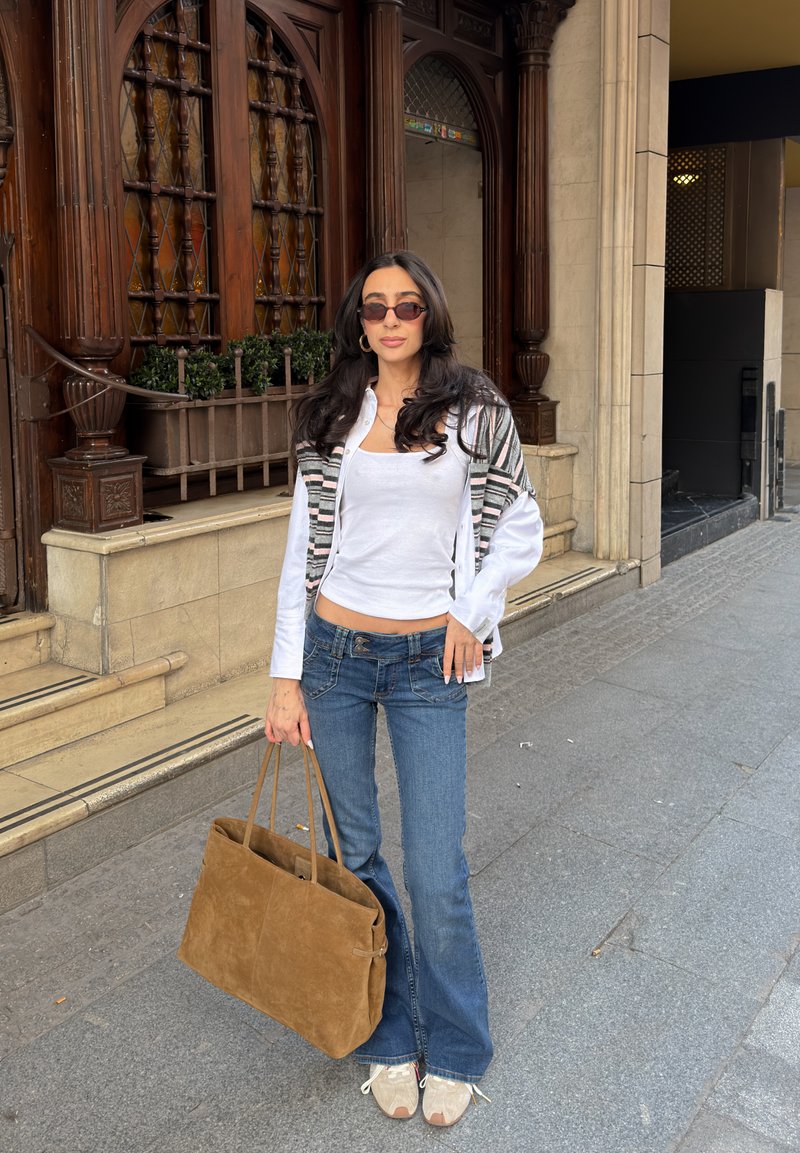 Γυναίκα με μακριά σκουρόχρωμα μαλλιά φοράει γυαλιά ηλίου, λευκό τοπ, ριγέ ζακέτα, μπλε τζιν, λευκά αθλητικά παπούτσια, κρατώντας μεγάλη καφέ τσάντα σε πεζοδρόμιο της πόλης.