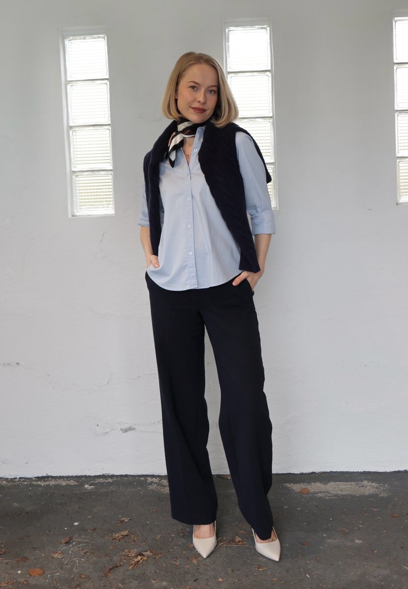 Femme portant une chemise bleu clair, une écharpe noire, un pantalon noir et des chaussures blanches, debout à l'intérieur contre un mur blanc avec trois fenêtres verticales.