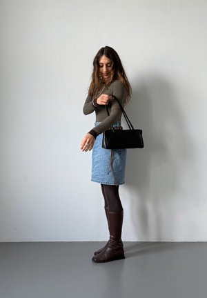 Mujer ajustando la correa de un bolso negro, vistiendo una blusa marrón de manga larga, falda vaquera, medias oscuras y botas marrones hasta la rodilla, de pie contra una pared blanca.