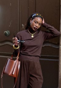 Femme en col roulé marron et pantalon tient des lunettes de soleil et un sac en cuir marron, appuyée contre une porte en bois sombre avec une main sur la tête.