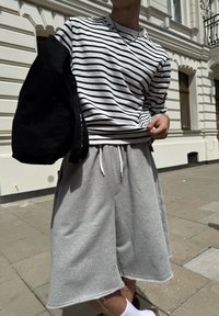 Prúžkovaná košeľa s dlhým rukávom v bielej a čiernej farbe, skombinovaná so sivými šortkami. Materiál vyzerá byť m�äkký, s viditeľnou šnúrkou a rozstrapkanými okrajmi.