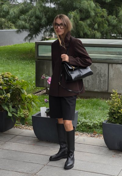 Botas de cuero negras hasta la rodilla, americana marrón a medida y pantalones cortos a medida. Los accesorios incluyen un bolso negro. Plantas verdes al fondo.