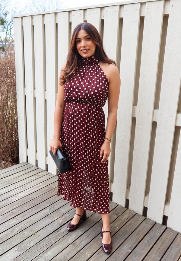 Μια γυναίκα στέκεται σε ξύλινη βεράντα φορώντας ένα αμάνικο μπορντό φόρεμα με λευκά πουά, κρατώντας ένα μαύρο τσαντάκι και φορώντας ταιριαστά παπούτσια.