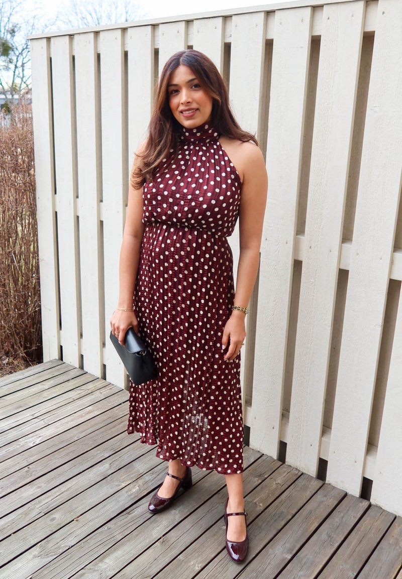 Une femme se tient sur une terrasse en bois, portant une robe sans manches bordeaux à pois blancs, tenant une pochette noire et portant des chaussures assorties.