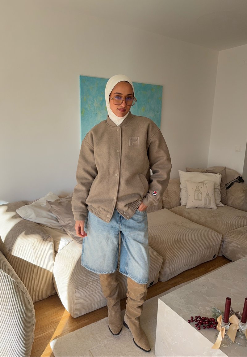 Veste polaire beige avec boutons-pression, jupe en denim bleu clair et bottes beige montant jusqu'aux genoux. La pièce comprend un canapé beige et des coussins décoratifs.