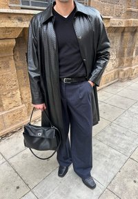Homme portant un manteau en cuir noir texturé, un pull noir, un pantalon bleu marine à rayures fines, des chaussures noires, tenant un sac à main en cuir noir sur un trottoir en ville.