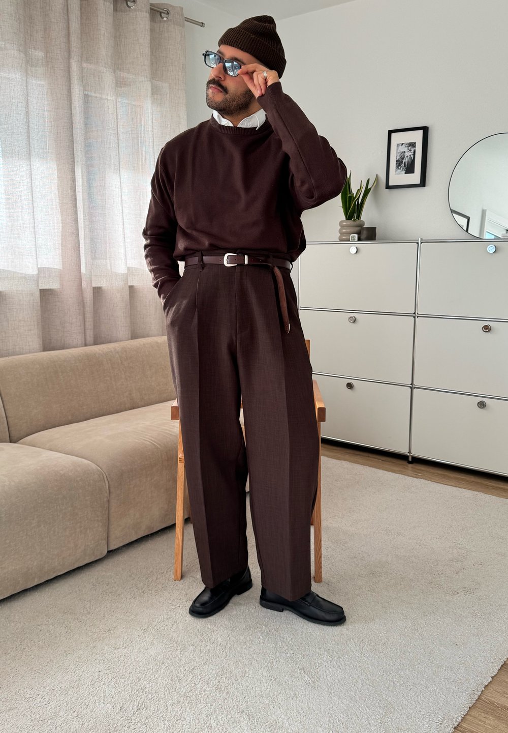 Pull tricot marron, pantalon marron taille haute ajusté avec plis, ceinture noire, chaussures noires et bonnet marron assorti. Éclairage intérieur doux.