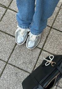 Srebrni škornji s sijočo površino, beli vezalke in zasnova tenisk. V kombinaciji z modrimi kavbojkami, na teksturirani asfaltni podlagi, poleg črne torbe.