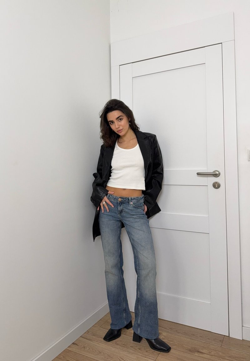 Schwarze Blazer, weißes kurz geschnittenes Tanktop, ausgebleichte blaue Schlagjeans und schwarze Stiefeletten mit Absatz. Glatte Texturen und lässiger Schnitt. Weiße Tür im Hintergrund.