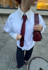 Camicia bianca con maniche a sbuffo, cravatta color marrone scuro, pantaloni larghi blu navy e borsa bordeaux. Gli accessori includono anelli e un orologio classico.