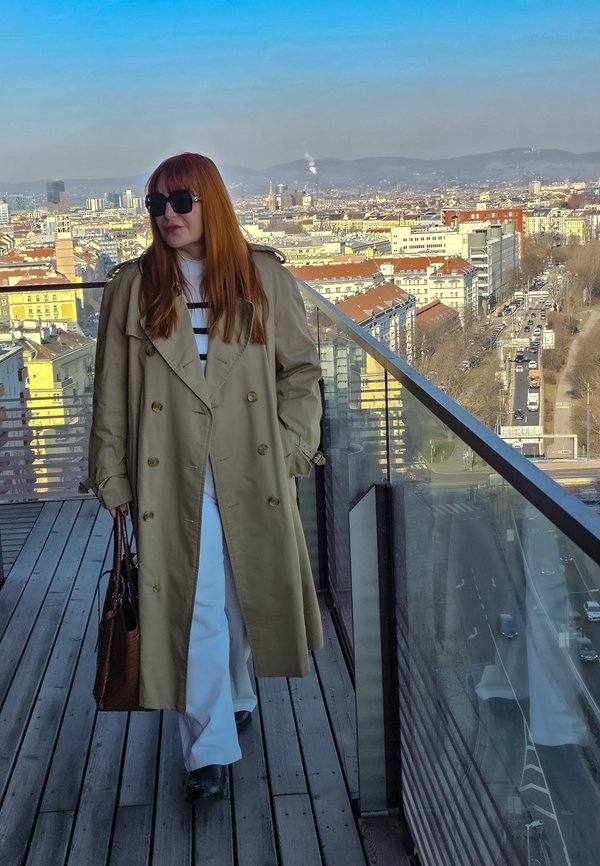 Vrouw met lang rood haar, beige trenchcoat, witte broek, zwarte zonnebril en een bruine tas, lopend op een balkon op het dak.