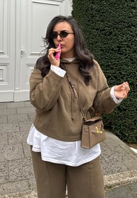 Kobieta w okularach przeciwsłonecznych i brązowym stroju nakłada różową pomadkę na szarym chodniku obok białych drzwi i zielonego żywopłotu.