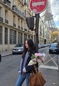 Nő rózsaszín bazsarózsákból álló csokorral, tengerészkék blézert és csíkos sálat viselve, egy utcatábla mellett, háttérben az Eiffel-torony.