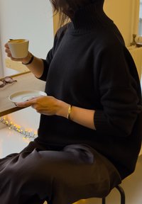 Pull noir à col roulé et pantalon foncé, tenant une tasse blanche avec une soucoupe, assis à une table en bois sous un éclairage ambiant chaleureux.
