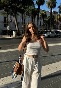 Top corto bianco e pantaloni bianchi larghi. Borsa marrone con una sciarpa blu e con un motivo floreale. Ambientazione urbana con palme.