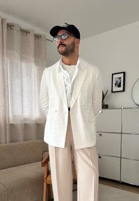 Férfi napszemüvegben, fekete sapkában, fehér blézerben és világos nadrágban, semleges tónusú nappaliban áll, mögötte kanapé és szekrény.