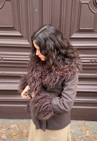 Brun jakke med teksturerte, lodne pelslignende detaljer på ermene, båret over et khakifarget skjørt. Bakgrunnen har detaljer fra en tredør.