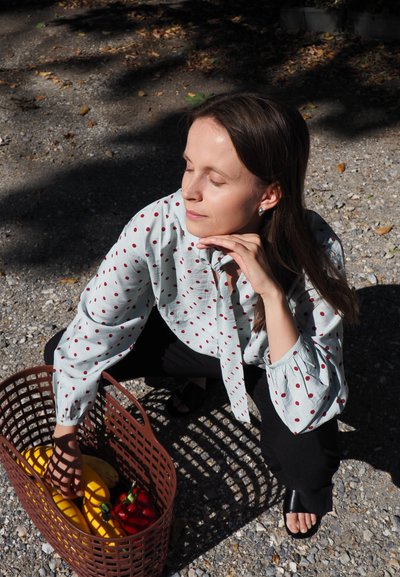 Camisa de color azul claro con lunares rojos, metida en pantalones negros. Sosteniendo una cesta marrón con plátanos y pimientos. Fondo de grava iluminado por el sol.