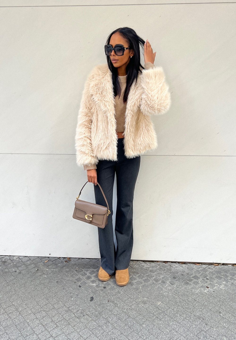 Chaqueta de piel sintética crema, suéter beige corto, pantalones grises ajustados, bolso marrón con cierre circular y botines marrón claro. Gafas de sol puestas.