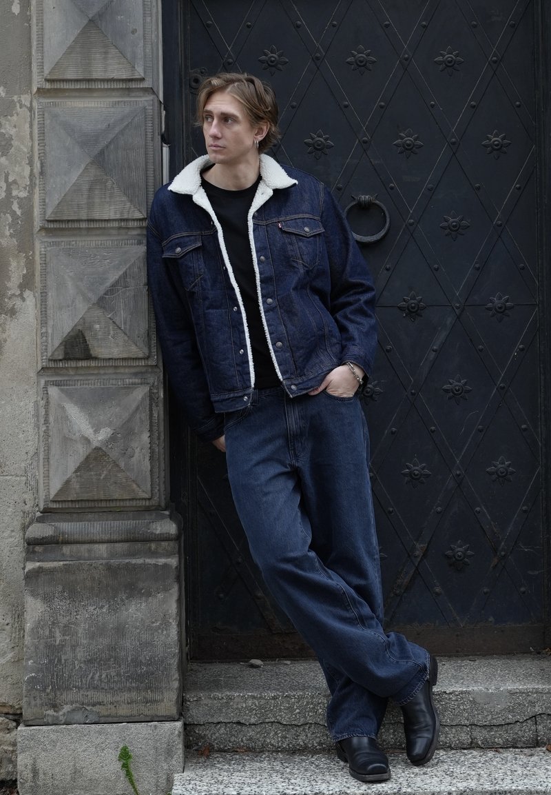 Jeune personne portant une veste en denim avec un col blanc et un jean, appuyée contre un mur de pierre à côté d'une porte métallique noire avec des clous décoratifs.