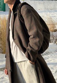 Personne portant un manteau à carreaux marron, un T-shirt blanc, un pantalon de survêtement Nike gris et un sac à dos noir, debout à l'extérieur sur un sol enneigé.