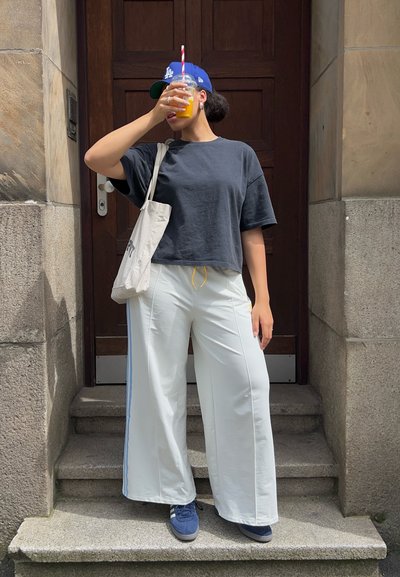 Camiseta negra corta, pantalones anchos blancos con rayas laterales, zapatillas azules y una gorra azul. Sosteniendo una bebida con una pajita de rayas.