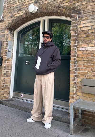 Sudadera con capucha gris oscuro y bolsillo canguro, pantalones anchos beige y zapatillas blancas, frente a una pared de ladrillo y una puerta verde.