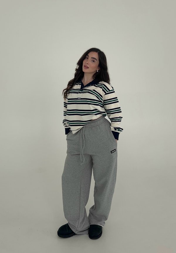 Femme debout, les mains dans les poches, portant un t-shirt rayé à manches longues, un pantalon de survêtement gris et des chaussures noires sur un fond uni.