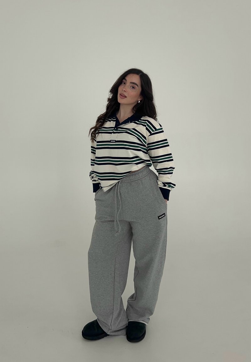 Frau steht mit Händen in den Taschen, trägt ein gestreiftes Langarmshirt, graue Jogginghose und schwarze Schuhe vor einfachem Hintergrund.
