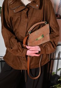 Brun mockaväska med gröna och gyllene detaljer, rektangulär form, avtagbar rem, hålls framför en brun jacka.