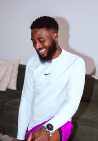 Homme souriant avec une barbe, portant un t-shirt blanc à manches longues Nike et un short violet, assis à l'intérieur près d'un canapé vert foncé avec des coussins.