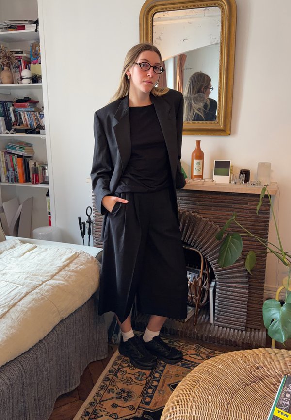 Black tailored blazer over a black top, wide-leg black trousers, and black shoes. Room with a retro mirror, bookshelves, and plants.