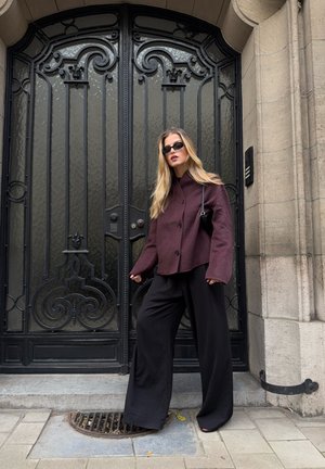 Chaqueta burdeos con cuello alto, pantalones de pierna ancha negros y un pequeño bolso negro. Textura suave, hardware mínimo, de pie frente a una puerta decorativa.