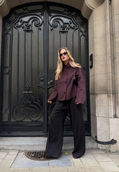 Chaqueta burdeos con cuello alto, pantalones de pierna ancha negros y un pequeño bolso negro. Textura suave, hardware mínimo, de pie frente a una puerta decorativa.