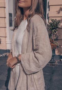 Ανοιχτό μπεζ λινό κασκόλ με φαρδιές μανίκες και χαλαρή εφαρμογή, φορεμένο πάνω από ένα ασημένιο υφαντό μπλουζάκι. Ο φορέας έχει έναν ασημένιο ρολόι στους καρπούς του.