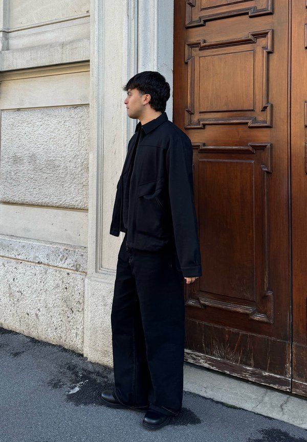 Personne portant une veste noire, un pantalon large noir et des bottes, debout de côté près d'un mur blanc texturé et d'une grande porte en bois marron.