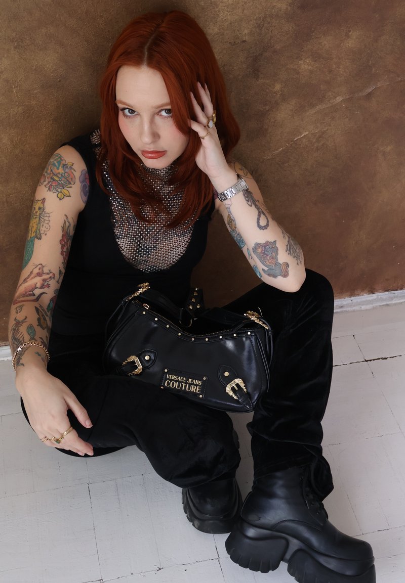 Femme aux cheveux roux avec des bras tatoués, assise par terre, vêtue d'une tenue noire et de bottes épaisses, tenant un sac à main noir Versace Jeans Couture.