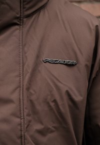 Brown insulated jacket with a smooth texture, zip closure, and a metallic logo detail on the left chest.