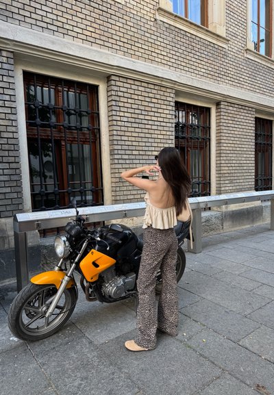 Motocicleta negra con acentos amarillos, con un faro redondeado y un motor plateado. Mujer con una blusa de volantes beige y pantalones estampados se encuentra al lado.