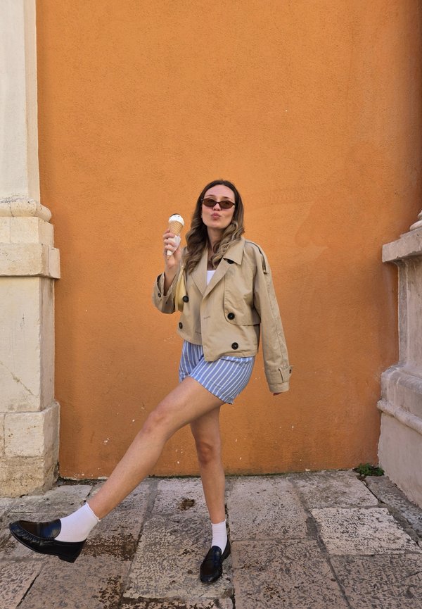 Veste beige courte avec des boutons croisés, shorts rayés bleus, mocassins noirs et chaussettes blanches, tenant un cornet de glace.