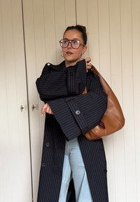 Čierny kabát s tenkými prúžkami, nadmerné rukávy, veľké gombíky a hnedá kožená kabelka na rameno. Svetlo modré džínsy dopĺňajú outfit.