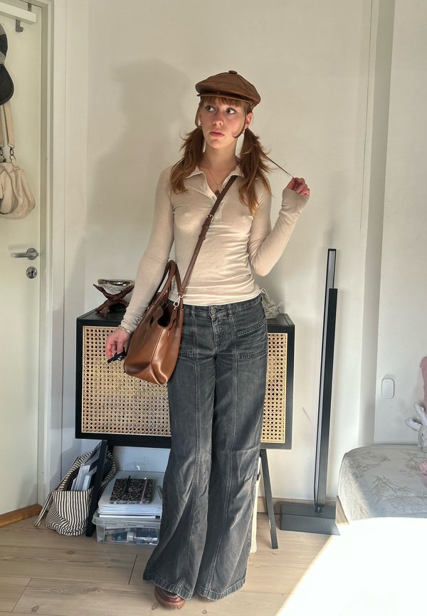 Una mujer posa en un interior con una gorra marrón, una camiseta color crema, unos vaqueros grises holgados y un bolso bandolera marrón.